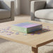 A 1000-piece jigsaw puzzle box sits on a light wood coffee table, surrounded by scattered puzzle pieces in a softly blurred living room setting with beige sofas in the background.
