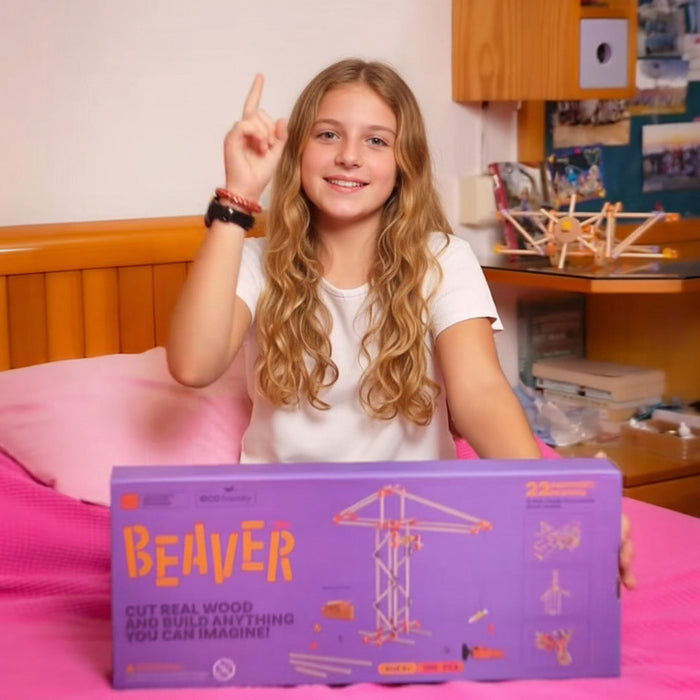 A smiling young girl with long light brown hair holds a large lavender box for a toy set in a bright, modern room. The box is branded 'Beaver' and features orange text and illustrations of wooden structures.