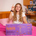 A smiling young girl with long light brown hair holds a large lavender box for a toy set in a bright, modern room. The box is branded 'Beaver' and features orange text and illustrations of wooden structures.