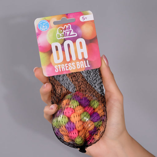 A hand holding a packaged stress ball in a mesh bag. The product is labeled 'Odd Ballz DNA Stress Ball' on a pink and orange header card, and the ball inside is multicolored with a black mesh exterior.