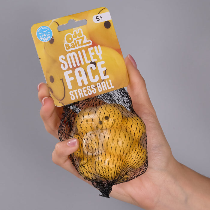 A hand holds a packaged yellow stress ball in a black mesh bag with a printed header card reading 'Odd Balz Smiley Face Stress Ball' against a plain gray background.