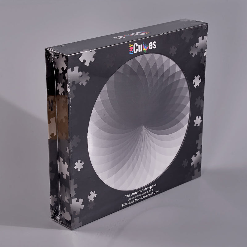 A boxed jigsaw puzzle package standing upright on a light gray studio surface. The box is dark gray/black with a large circular white-to-gray puzzle design on the front, scattered white puzzle-piece silhouettes around the edges, and the visible brand name 'Cubes' near the top.