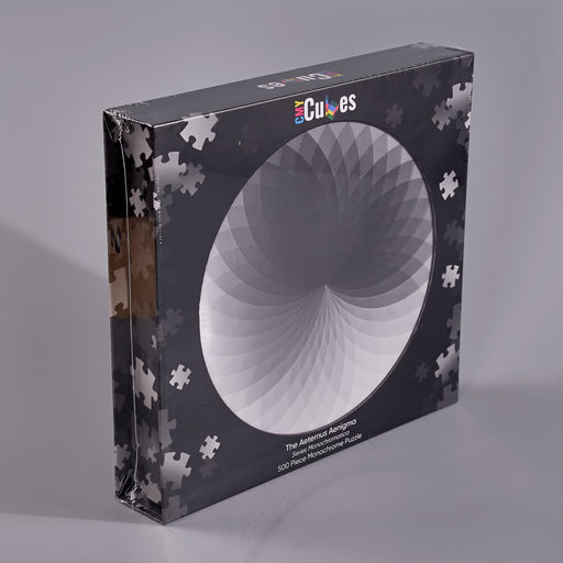 A boxed jigsaw puzzle package standing upright on a light gray studio surface. The box is dark gray/black with a large circular white-to-gray puzzle design on the front, scattered white puzzle-piece silhouettes around the edges, and the visible brand name 'Cubes' near the top.