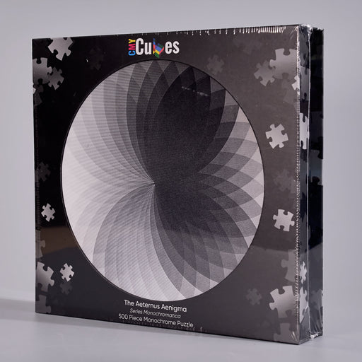 A boxed jigsaw puzzle package on a light gray studio background. The front shows a circular black-and-white optical illusion design with the brand name 'Cubes' at the top and text reading 'The Asterus Aenigma 500 Pieces Monochrome Puzzle'.