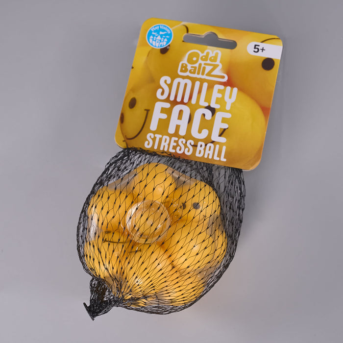 Odd Ballz Smiley Face Stress Ball in a black mesh bag with a yellow cardboard hanger, photographed on a neutral gray background.