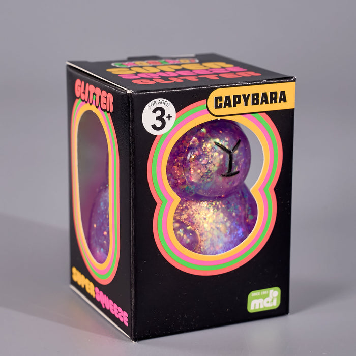 A boxed toy product on a plain gray studio background. glitter capybara combines soft, tactile comfort with shimmering sparkle that catches the light beautifully. The black retail box shows a window revealing two glittery iridescent purple spherical toys with rainbow concentric rings printed on the package.