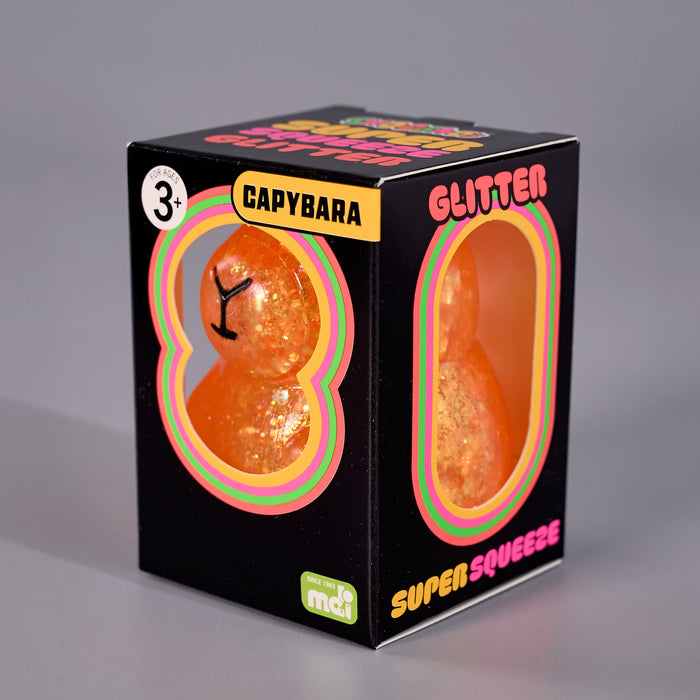 A black retail box for a Capybara-shaped glitter squishy toy, with two oval cutouts showing the orange glittery toy inside. The box has colorful text including 'GLITTER', 'SUPER SQUEEZE', 'Capybara', and a small 'mci' logo, photographed on a neutral gray background.