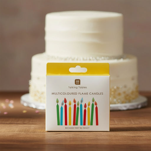 A boxed set of multicolored flame candles from Talking Tables stands on a wooden surface in front of a blurred two-tier white cake. The package is white with a yellow top panel and an illustration of colorful candles.