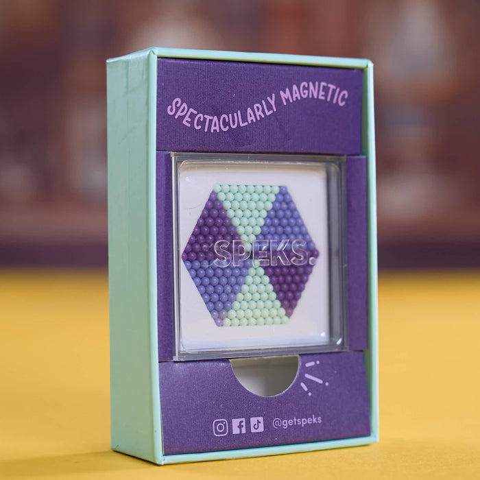 A boxed magnetic bead puzzle on a yellow surface with a blurred brown background. The mint green and purple packaging shows a hexagonal bead arrangement in purple, blue, and mint green, with the text 'SPECTACULARLY MAGNETIC' and the brand name 'SPEKS.