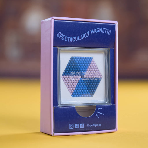 A boxed product labeled 'SPECTACULARLY MAGNETIC' standing upright on a yellow surface with a blurred brown background. The package is pink and dark blue, with a clear window showing a hexagonal magnetic bead arrangement in blue and pink.