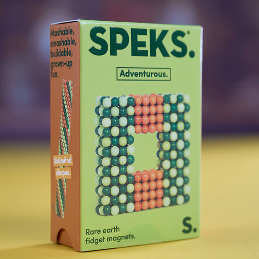 A box of SPEKS. fidget magnets, featuring a light green front with a colorful magnetic bead design and an orange side panel with product text, set against a blurred yellow and purple background.