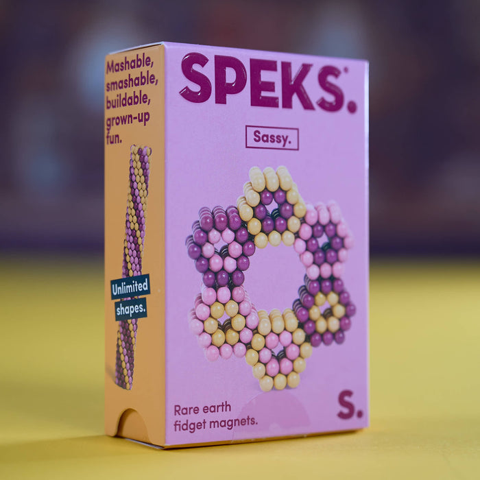 A pink and orange box of SPEKS. Sassy. fidget magnets, standing upright on a yellow surface with a blurred dark background.
