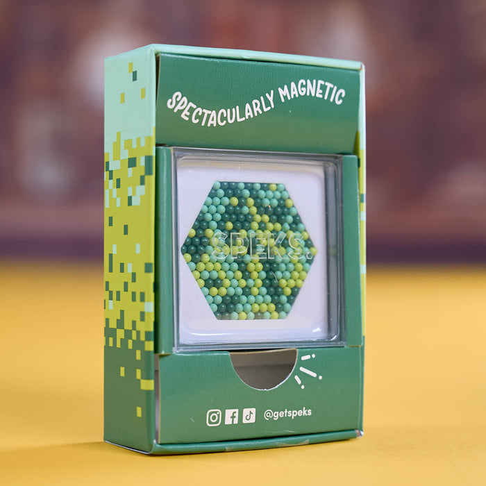 A green product box labeled 'SPECTACULARLY MAGNETIC' with a clear window showing a hexagonal cluster of small magnetic beads in green and yellow, photographed on a yellow surface with a blurred brown background.
