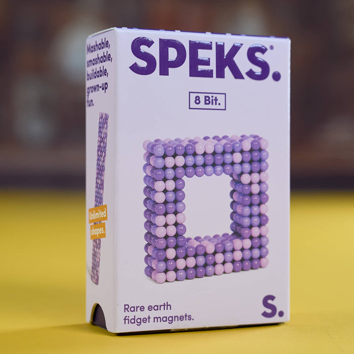 A box of SPEKS. 8 Bit rare earth fidget magnets, featuring a white box with purple branding and an image of a square frame built from purple and lavender magnetic beads, set against a yellow surface and blurred brown background.