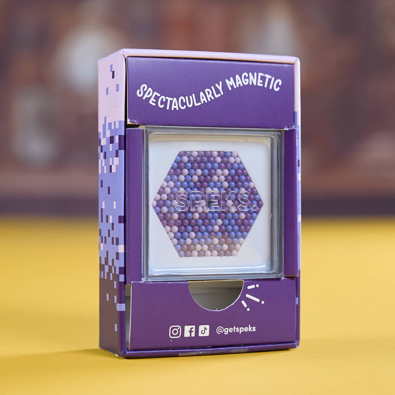 A purple product box labeled 'SPECTACULARLY MAGNETIC' with a clear window showing a hexagonal arrangement of small magnetic beads. The box has pixel-style side graphics and social media icons near the bottom, standing on a yellow surface against a blurred brown background.