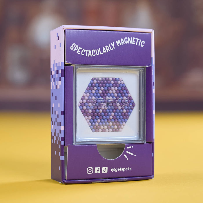 A purple product box labeled 'SPECTACULARLY MAGNETIC' with a clear window showing a hexagonal arrangement of small magnetic beads. The box has pixel-style side graphics and social media icons near the bottom, standing on a yellow surface against a blurred brown background.