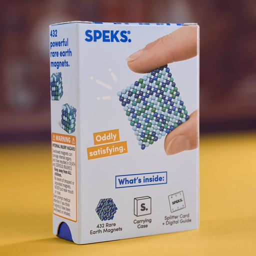 Speks Rare Earth Magnets kit box, showing 432 powerful rare earth magnets in a blue and green beaded square design, held by a hand on a yellow surface with a blurred brown background.