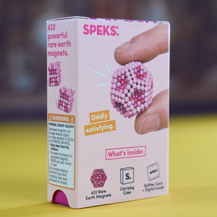 A product box for 'Speks Rare Earth Magnets' toy, showing a hand holding a small cluster of pink and white magnetic beads. The box is white with pink and orange text, set against a yellow surface and a blurred dark background.
