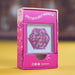 A pink retail box for a 'Spectacularly Magnetic' toy, featuring a hexagonal window displaying colorful magnetic beads. The box has pixelated side patterns and social media handles at the bottom, set against a yellow surface with a blurred background.