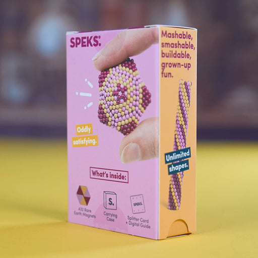 A pink and orange box of SPEKS. bead art kits, featuring a hand holding a beaded heart-shaped piece on the front, set against a blurred background with a yellow surface.