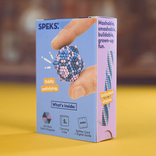 A Speks bead-making kit box standing upright on a yellow surface with a blurred brown background. The box is light blue and lavender, showing a hand holding a blue and pink beaded ball, with text including 'SPEKS.