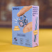 A Speks bead-making kit box standing upright on a yellow surface with a blurred brown background. The box is light blue and lavender, showing a hand holding a blue and pink beaded ball, with text including 'SPEKS.