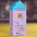 A house-shaped cardboard box for kids DIY perfume, featuring a light purple body with a bright blue top, cartoon graphics, and step-by-step instructions on how to make the perfume. The box is photographed standing upright on a yellow surface with a blurred brown background.