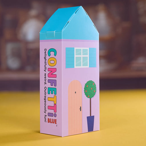 A colorful house-shaped box for Confetti Blue confetti, standing on a yellow surface with a blurred brown background. The box features a light purple body, a bright blue roof, a peach door, and a green tree illustration.