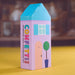 A colorful house-shaped box for Confetti Blue confetti, standing on a yellow surface with a blurred brown background. The box features a light purple body, a bright blue roof, a peach door, and a green tree illustration.