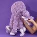 Purple plush octopus, soft fluffy texture, lavender color, cute smiling face, long tentacles, white rope-detail accents, cuddly stuffed toy, raised display, close-up product focus.