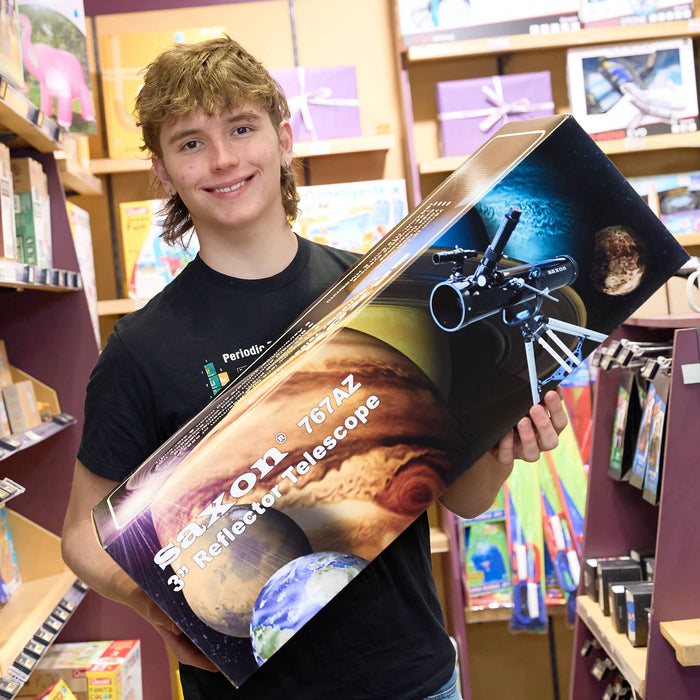 A smiling young man in a black t-shirt holds a large Saxon 76mm 3 Reflector Telescope box inside what appears to be a toy or science store.