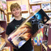 A smiling young man in a black t-shirt holds a large Saxon 76mm 3 Reflector Telescope box inside what appears to be a toy or science store.