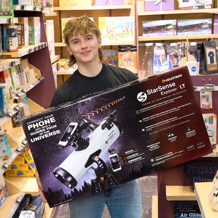 A young man holding a very large Celestron StarSense Explore LT telescope box inside a toy or hobby store.