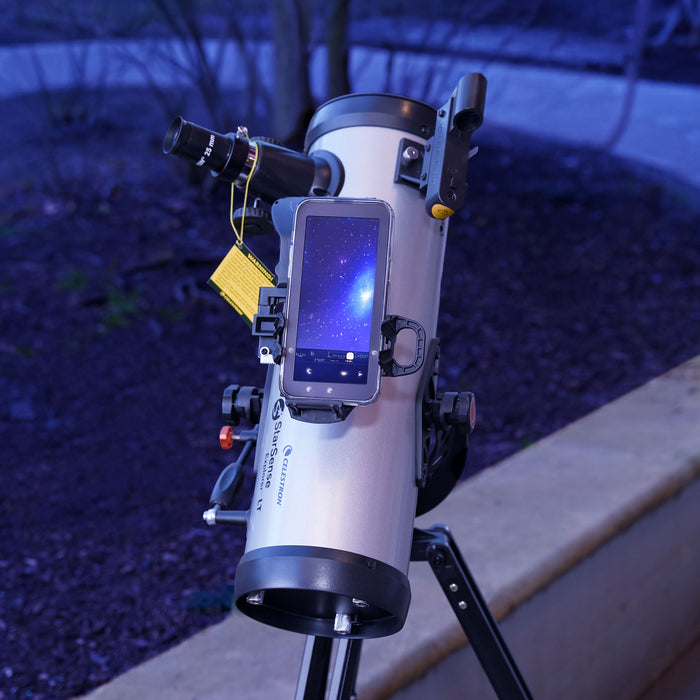 A white and black Newtonian-style telescope mounted on a tripod, with a smartphone attached to the side displaying a starry night sky. The scene is photographed outdoors at night with a blurred dark ground and light-colored concrete curb in the background.