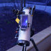 A white and black Newtonian-style telescope mounted on a tripod, with a smartphone attached to the side displaying a starry night sky. The scene is photographed outdoors at night with a blurred dark ground and light-colored concrete curb in the background.