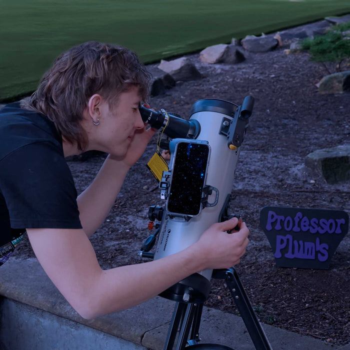 A person outdoors looking through a telescope mounted on a tripod, with a smartphone attached to the tube. A sign reading 'Professor Plum's' is visible on the ground nearby.