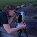 A person outdoors looking through a telescope mounted on a tripod, with a smartphone attached to the tube. A sign reading 'Professor Plum's' is visible on the ground nearby.
