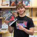 A young man in a black graphic t-shirt stands in a toy or hobby store, holding a book titled 'The Power Up Paper Airplane Book' and pointing to a periodic table graphic on the shirt.