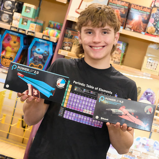A smiling young man in a black t-shirt holds two boxed products in a retail store setting. The boxes are branded POWER UP 2.