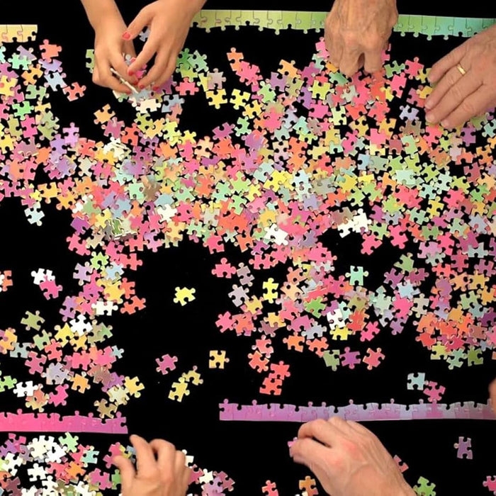 A large assortment of colorful jigsaw puzzle pieces scattered across a black surface, with multiple hands visible at the edges sorting or holding the pieces.