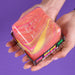 A person's hands holding a square plastic container filled with a marbled, translucent pink, orange, and yellow substance against a solid purple background, 'Nice Berg Swirl'