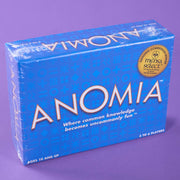 A blue box of Anomia card game, sealed in clear plastic wrap, set against a solid purple background. The box features the game's title in large white letters, along with a gold Mensa Select stamp.