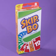 A boxed card game called Skip-Bo, photographed upright against a solid purple background. The box is mostly white with large orange and blue Skip-Bo lettering, plus green and yellow sections and sample card graphics showing red, green, blue, and orange cards.