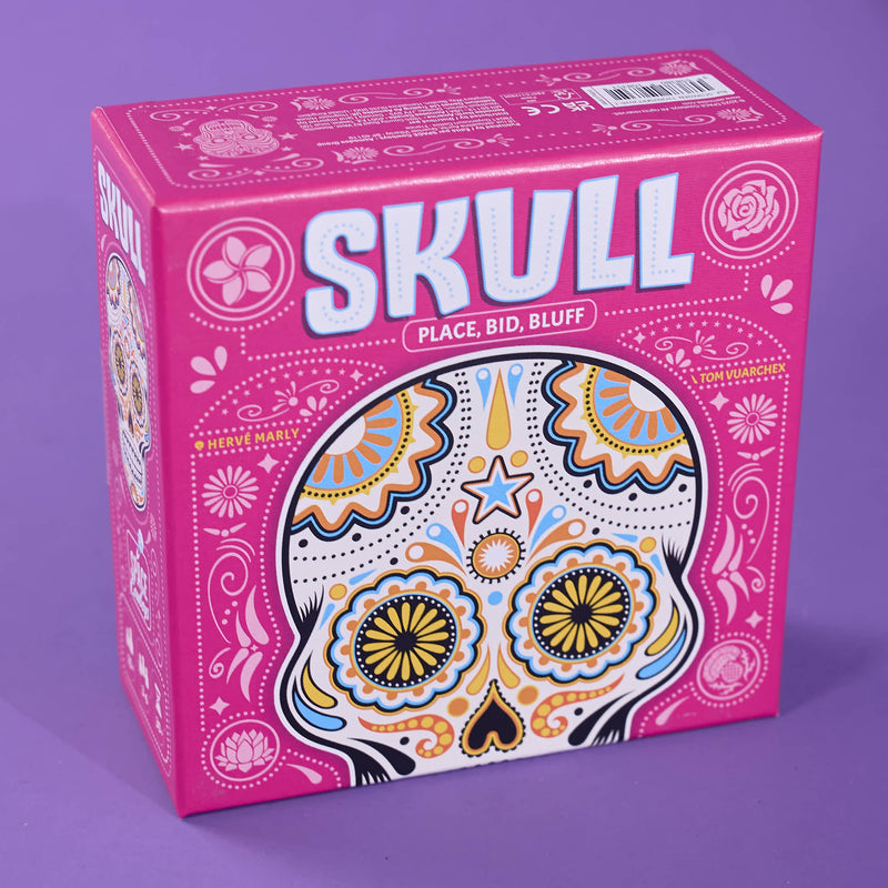 The product packaging for the card game 'Skull: Place, Bid, Bluff' is shown on a solid purple background. The box is bright pink with white text and a colorful sugar skull illustration on the front.