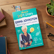 A children's book titled 'Aussie STEM Stars Emma Johnston' lies on a wooden tabletop, surrounded by a pencil, glasses, a small plant, and other books. The cover features a bright teal background with an illustrated woman in a diving suit and marine-themed graphics.