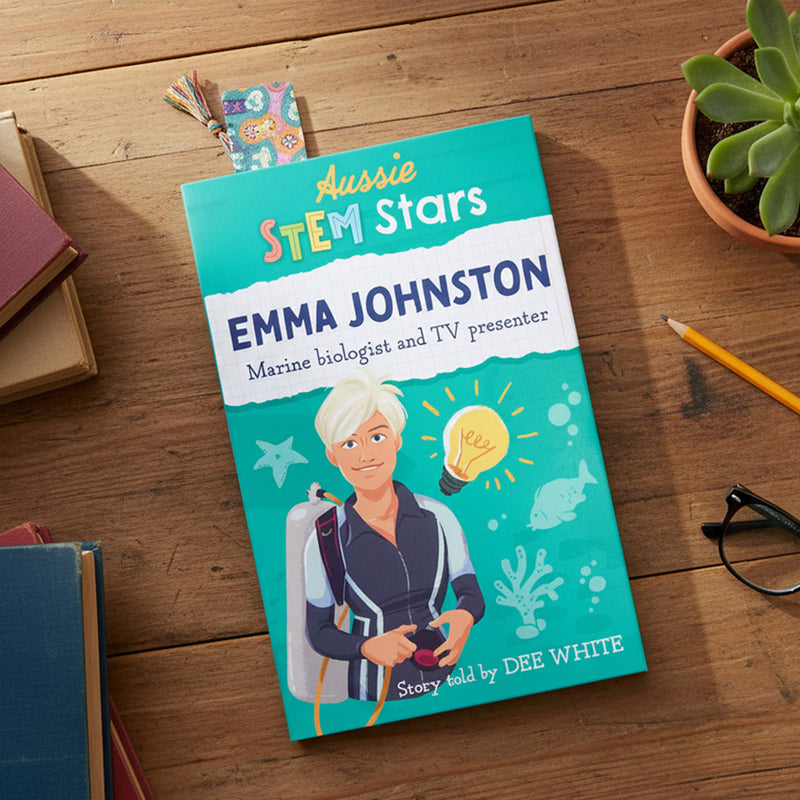 A children's book titled 'Aussie STEM Stars Emma Johnston' lies on a wooden tabletop, surrounded by a pencil, glasses, a small plant, and other books. The cover features a bright teal background with an illustrated woman in a diving suit and marine-themed graphics.