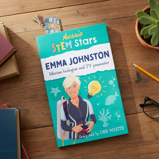 A children's book titled 'Aussie STEM Stars Emma Johnston' lies on a wooden tabletop, surrounded by a pencil, glasses, a small plant, and other books. The cover features a bright teal background with an illustrated woman in a diving suit and marine-themed graphics.