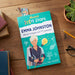 A children's book titled 'Aussie STEM Stars Emma Johnston' lies on a wooden tabletop, surrounded by a pencil, glasses, a small plant, and other books. The cover features a bright teal background with an illustrated woman in a diving suit and marine-themed graphics.