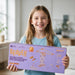 A smiling young girl with long light brown hair holds a large lavender box for a toy set in a bright, modern room. The box is branded 'Beaver' and features orange text and illustrations of wooden structures.