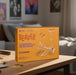 A yellow-orange product box labeled 'BEAVER' sits on a wooden table in a softly blurred bedroom or playroom setting. The box shows a wooden airplane model and text including 'CUT REAL WOOD AND BUILD ANYTHING YOU CAN IMAGINE!' and '10 CRAFTERS, 1000+ PIECES.